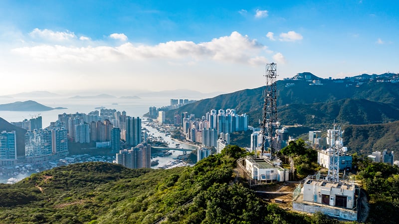 Aerial view of a city with tall buildings along a waterway, surrounded by green hills. A communication tower supporting mobile-enabled early warning systems sits atop a hill in the foreground. Blue sky, scattered clouds, and distant mountains complete the scene.