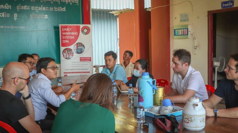 A group of people sit around a wooden table in a meeting room, discussing. Some wear glasses; one person wears a mask. On the table are thermoses, cups, and bags. A poster about mobile-enabled early warning systems is displayed behind them.