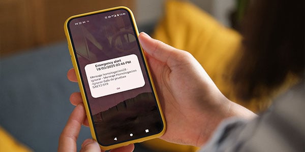 A person holding a smartphone with a yellow case receives an urgent notification from Chile's Emergency Alert System. The alert on the screen shows a date, time, message, and "Ok" button. A yellow pillow and part of a gray sleeve appear in the background.