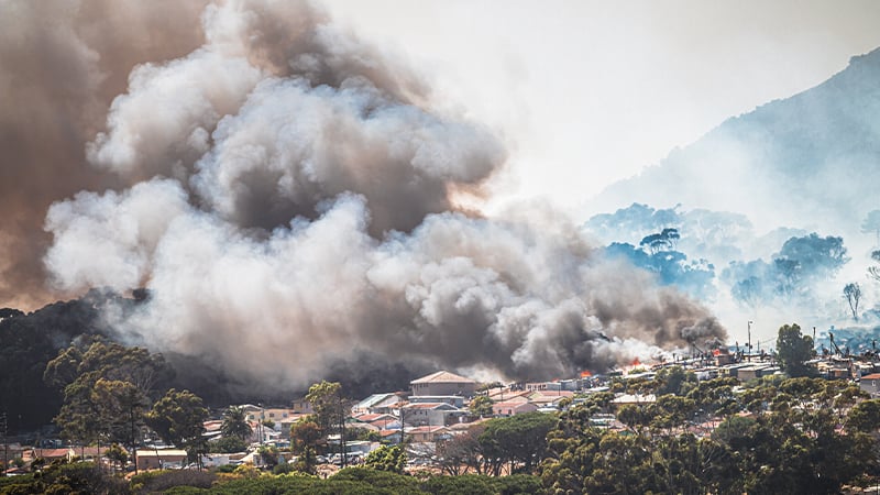 Thick gray and black smoke billows above a residential neighborhood as a wildfire burns nearby, obscuring houses and trees. In this densely packed area, mobile-enabled early warning systems are crucial for alerting residents to danger.