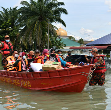 A rescue worker guides a red boat carrying people in life jackets and masks, along with bags and boxes, through flooded streets—a striking example of humanitarian innovation amid crisis. Palm trees and buildings stand under a clear sky in the background.