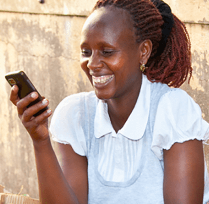 A smiling woman with braided hair in a black scrunchie looks at a smartphone, possibly exploring GSMA Mobile for Development. She wears a white blouse with a light blue vest and gold earrings, sitting outdoors against a sunlit beige concrete wall.