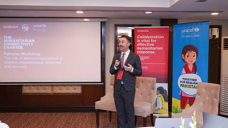 A man in a suit speaks into a microphone at a workshop. Behind him are banners from GSMA and UNICEF, plus a large screen displaying “The Humanitarian Connectivity Charter” and info on how Pakistan can respond to and recover from disasters using telecommunications. Two empty chairs nearby.