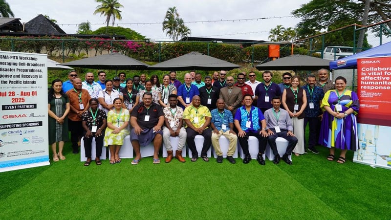 A group of about 30 people pose outdoors on green grass, some seated and others standing, with umbrellas and palm trees in the background. Event banners on the left detail dates and sponsors for a Pacific Islands disaster preparedness workshop to respond to sudden onset emergencies.