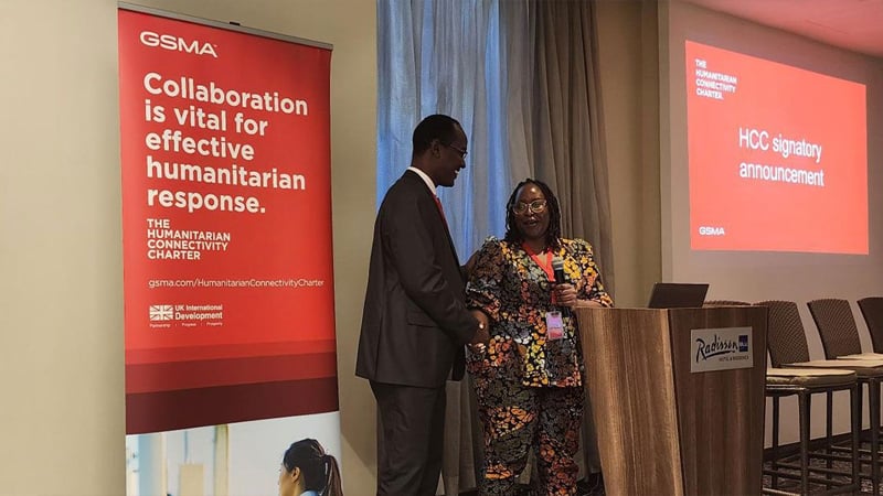 Two people stand and talk at a podium beside a large screen and banner with red backgrounds, highlighting the Humanitarian Connectivity Charter. The banner notes, “Collaboration is vital to prepare for and respond to crises,” at this GSMA event in Radisson Blu.