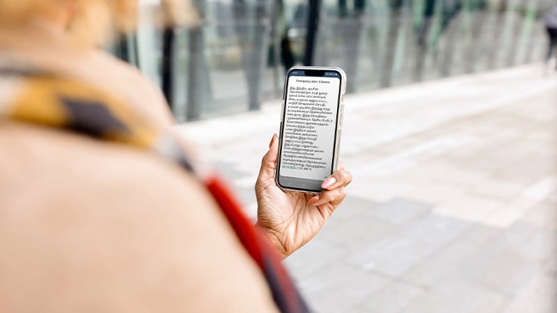 A person holds a smartphone outdoors, reading emergency care information powered by mobile-enabled early warning systems. The background features a modern glass building and paved sidewalk, with the phone in sharp focus and surroundings slightly blurred.