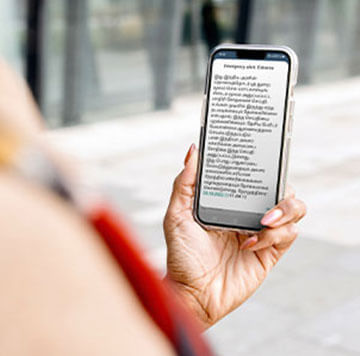 A person holds a smartphone in their left hand, reading a message in Tamil on the screen—showcasing how digital humanitarian assistance empowers communities. The background features a glass building and paved walkway.