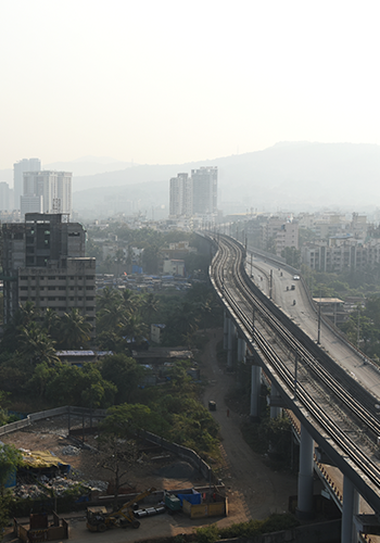 A hazy cityscape shows an elevated metro rail track curving through urban buildings and greenery. High-rise apartments and distant hills are visible under a pale sky, with a few vehicles on the road, creating a calm, early morning ambiance.