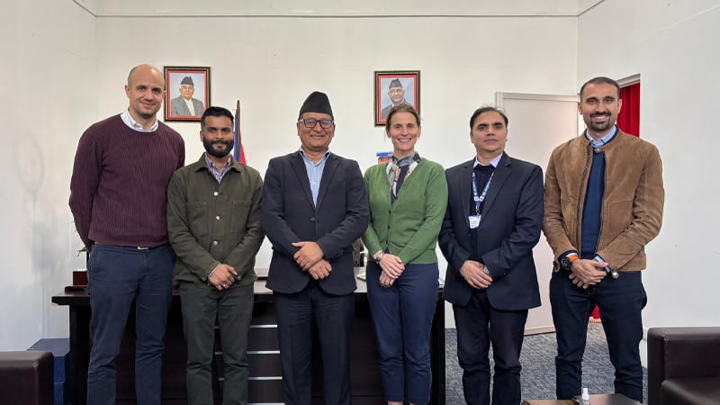 Six people stand in a row indoors, smiling at the camera. Among the group of four men and two women—experts in mobile-enabled early warning systems—are framed portraits, a Nepali flag, a desk, and a white wall behind them.
