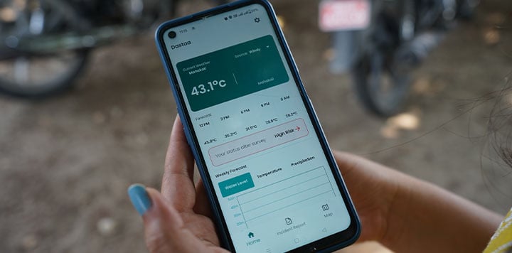 A hand holds a smartphone displaying a weather app, highlighting digital humanitarian assistance. The screen shows 43.1°C with detailed data, graphs, and weather options. In the blurred background, motorcycles and outdoor scenery are visible.