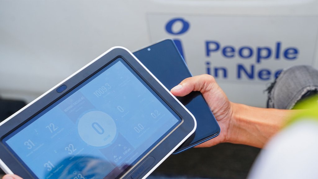 A person holds a tablet displaying data and a smartphone, with a blurred “People in Need” sign in the background. The scene highlights technology’s role in humanitarian work or data collection for anticipatory action in the Philippines.