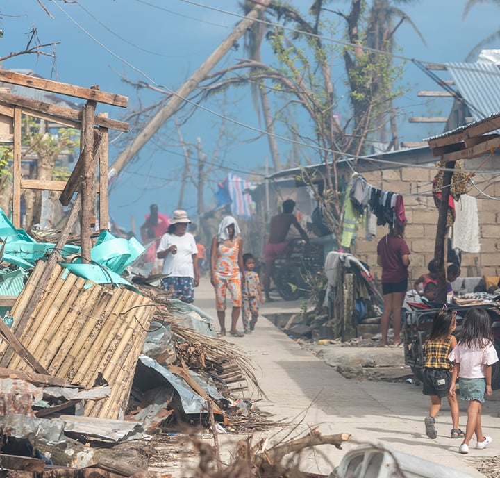 A group of people, including children, walk along a debris-strewn street between damaged homes. Fallen trees and rubble show recent devastation, highlighting the need for digital humanitarian assistance in the aftermath. The sky is blue and hazy.