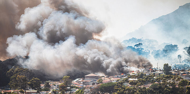Thick, dark smoke billows from a wildfire near homes surrounded by trees and hills. As flames threaten residences, digital humanitarian assistance plays a crucial role in coordinating relief efforts amid the hazy sky and faint mountains.