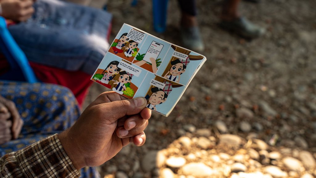A hand holds a small, colorful comic booklet with cartoon characters speaking in speech bubbles about blockchain for anticipatory action. In the background, blurred people sit on blue chairs outdoors on dirt and rocks, sunlight casting shadows.