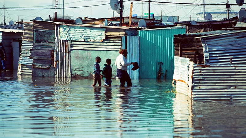 A woman and two children wade through knee-deep floodwater between makeshift corrugated metal shacks in an informal settlement, where mobile-enabled early warning systems could help residents prepare for rising waters. Satellite dishes dot several rooftops.