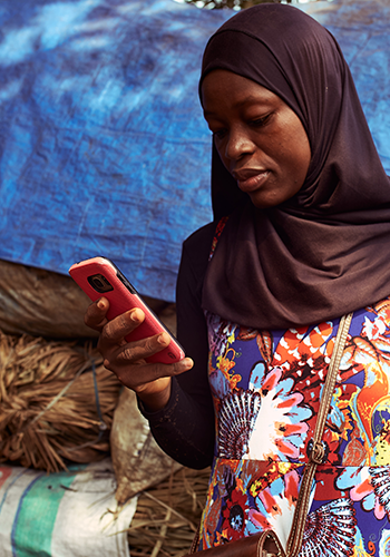 A woman wearing a black hijab and a colorful patterned dress looks down at her pink smartphone. She stands outdoors near what appears to be stacks of straw and a blue tarp, with a brown shoulder bag visible.
