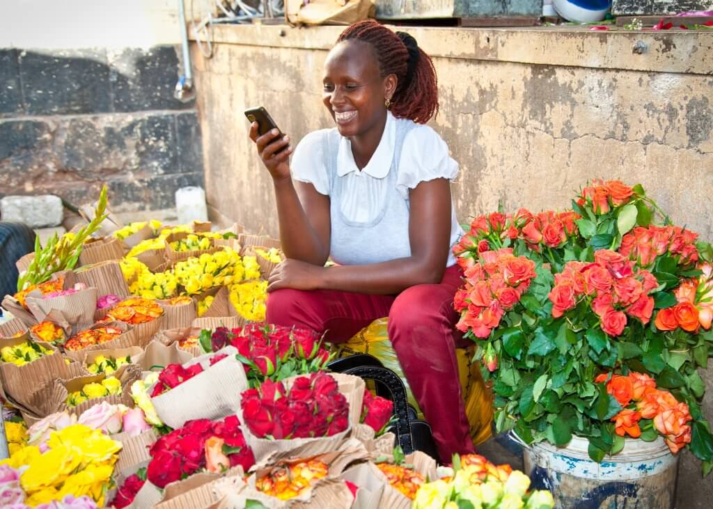 A smiling woman sits among colorful bouquets—yellow, red, orange, and pink—wrapped in brown paper at her market stall. In a white shirt and red pants, she looks at her smartphone, possibly managing sales through a mobile money API amid her floral arrangements.