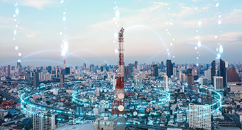 A red and white communication tower stands in the centre of a modern cityscape. Digital blue lines and dots arc between towers and buildings, symbolising wireless connectivity and data transfer. The sky is clear with soft clouds.