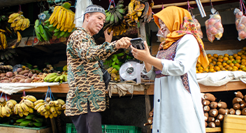 A man in patterned clothing pays a woman in a yellow headscarf with a card machine at an outdoor fruit market, surrounded by bananas, oranges, sweet potatoes, and other produce displayed on tables. Both are smiling and interacting warmly.