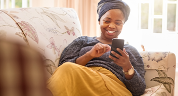 A smiling woman in a headwrap and casual clothes relaxes on a floral-patterned sofa, looking at her mobile and tapping the screen. Sunlight streams through a window in the background, creating a warm, cheerful atmosphere.