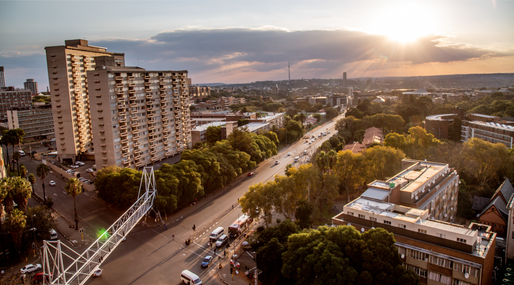 Aerial view of a city at sunset, with tall apartment buildings, tree-lined streets, and cars on the roads. The sun is low in the sky, casting golden light and rays through scattered clouds above the urban landscape.