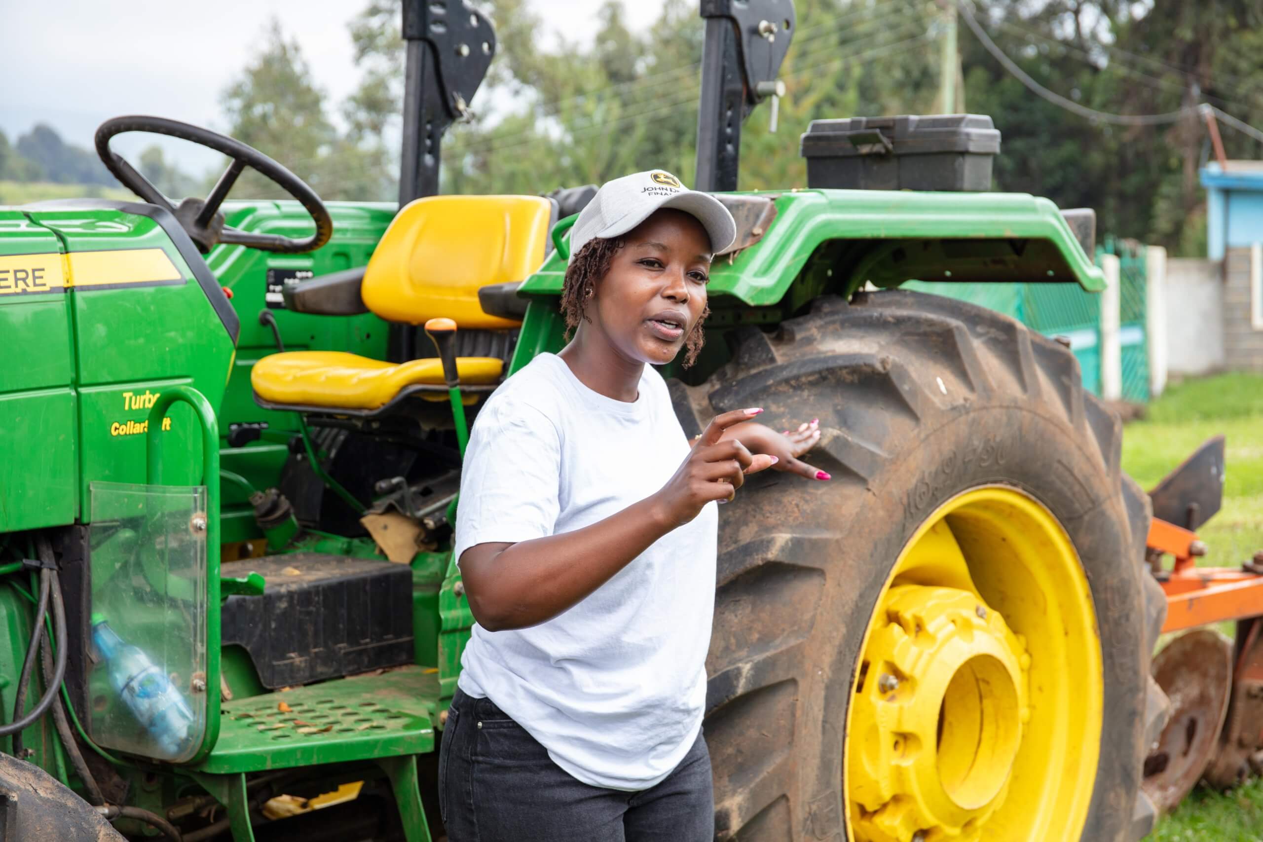 A woman in a white shirt and gray cap speaks while standing next to a large green and yellow tractor outdoors. She gestures with her hands, and the tractor’s steering wheel and big rear tire are clearly visible behind her. Trees and a building are in the background.