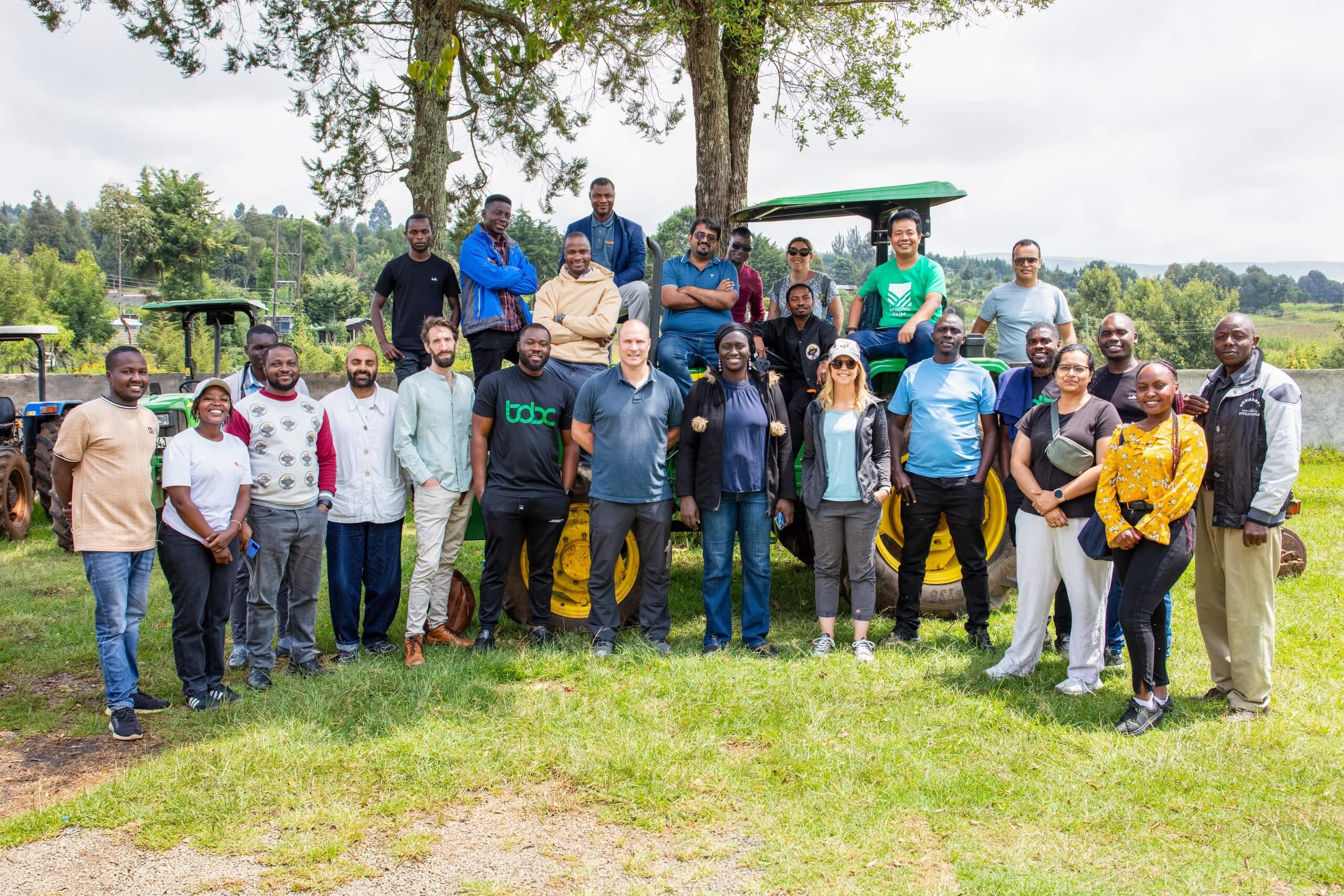 A diverse group of about 25 people stand and sit outdoors in front of a tractor, smiling at the camera. They are on grass with trees and farming equipment in the background, dressed in casual clothing, appearing friendly and relaxed.