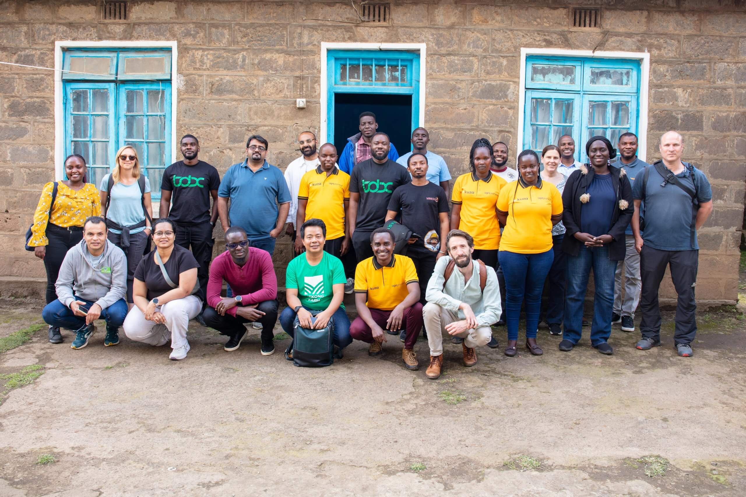 A diverse group of about 25 people, some wearing yellow shirts, pose and smile in front of a stone building with blue-trimmed windows and door. Some are standing, others are kneeling or sitting in front, and the background shows the building’s façade.
