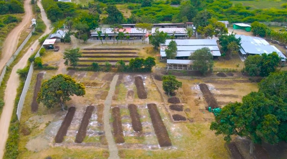 Aerial view of a rural farming area with rectangular garden beds arranged in rows. Paths run between the beds. Trees and green bushes surround the site. Buildings with metal roofs line the upper part, and two dirt roads border the area on the left.