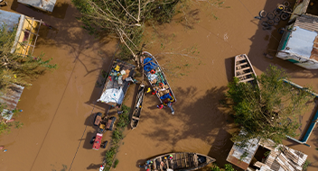 Aerial view of a flooded area with muddy water covering the ground. People in two boats navigate between partially submerged houses and trees. Some roofs and belongings are visible above water, showing damage and disarray from the flood.
