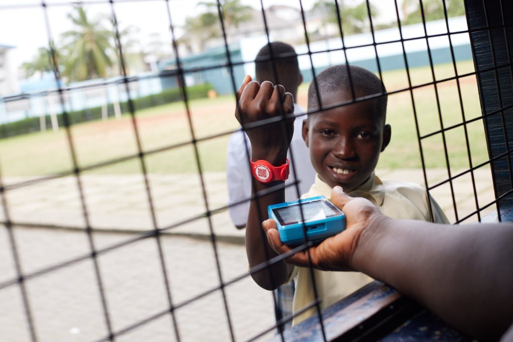 A smiling boy holds up his wrist with a red digital wristband toward a person scanning it through a metal fence. Another child stands nearby. The setting appears to be an outdoor sports area with palm trees and buildings in the background.