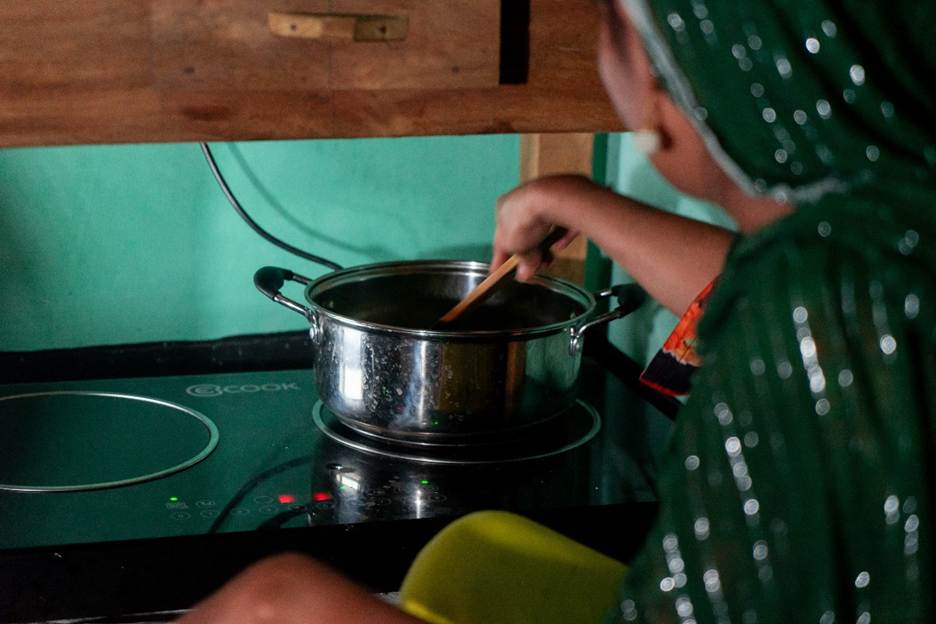 A person in a green headscarf stirs food in a stainless steel pot on an electric stove. The background has a wooden cabinet and teal-green wall. The person is viewed from behind and partially out of focus, with their right hand holding a wooden spoon.