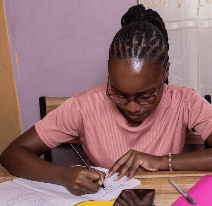 A young woman with braided hair and glasses sits at a wooden desk, studying for her mobile money certification. She writes in a notebook, surrounded by papers, a smartphone, and a pink folder. Behind her is a lavender wall and a window with a white curtain.
