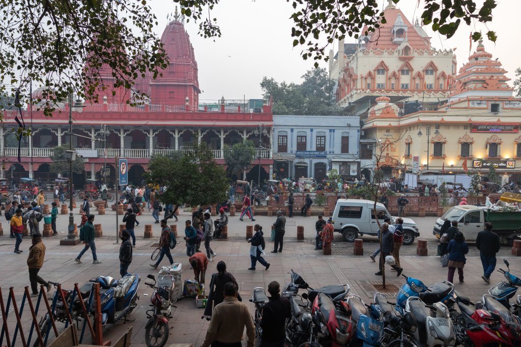 A busy street scene in India shows people walking and gathering near temples with ornate red and cream towers. Bikes and motorbikes are parked at the front, while trees frame the top of the image. Some cars and rickshaws are on the road.