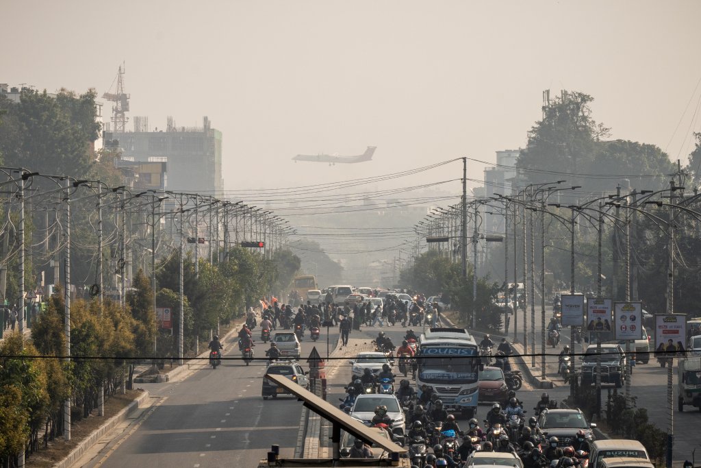 A busy city street is filled with vehicles and motorcycles in traffic. Overhead, power lines crisscross the scene. In the background, an airplane approaches for landing, partially obscured by hazy, smoggy air. Trees and buildings line the road.