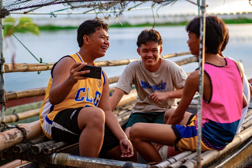 Three teenage boys sit on a bamboo raft, smiling and laughing together. One holds a smartphone for a selfie. The scene is outdoors by the water, lit by warm sunlight. They wear casual clothes, including sports jerseys and shorts.