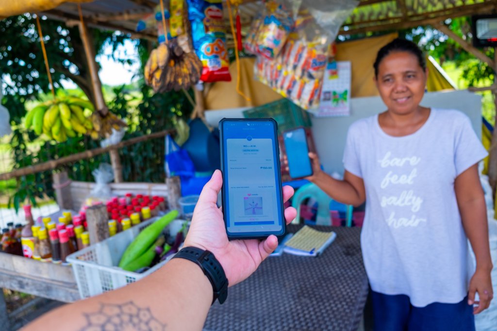 A person holds a mobile phone displaying a payment app in front of a smiling woman at an outdoor market stall selling fruit, sauces, and snacks. The stall is shaded and surrounded by greenery. The woman wears a light T-shirt and stands behind the counter.
