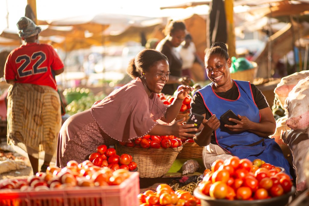Hero image for the COP30 campaign: Two women sit at a market stall, smiling and laughing as they look at a smartphone together. They are surrounded by baskets and crates filled with ripe red tomatoes. Other vendors and market stalls are visible in the sunny, bustling background.