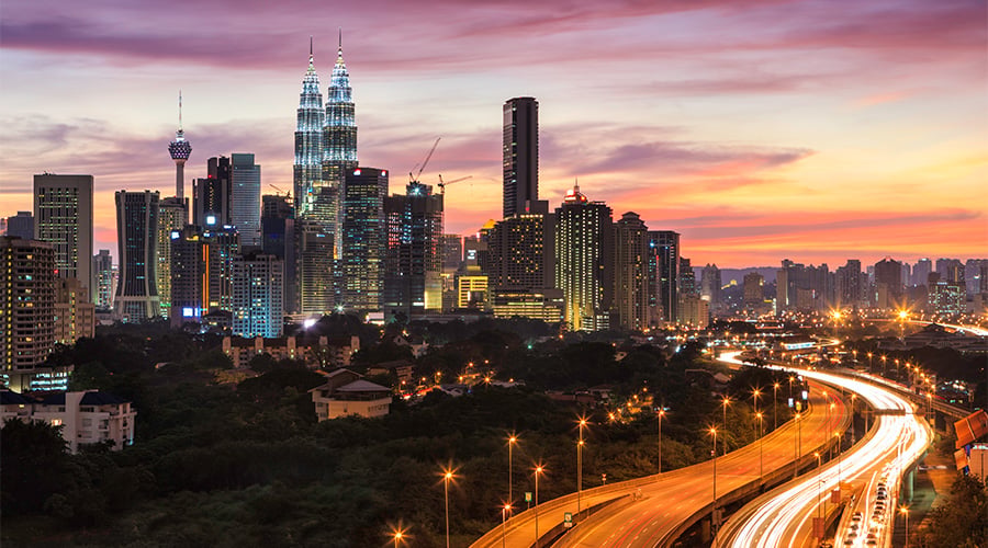 A vibrant cityscape at sunset shows Kuala Lumpur’s skyline, highlighting the illuminated Petronas Twin Towers and other skyscrapers. A curving motorway in the foreground has streaks of car lights, while the sky glows with pink, orange, and purple hues.