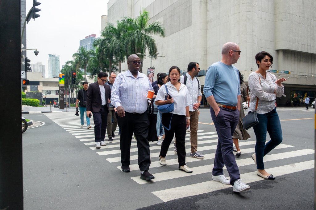 A diverse group of people in business and casual attire cross a city street at a pedestrian crossing. Some hold coffee cups, mobile phones, or bags. Tall buildings, palm trees, and traffic lights are visible in the background. The weather appears clear and mild.