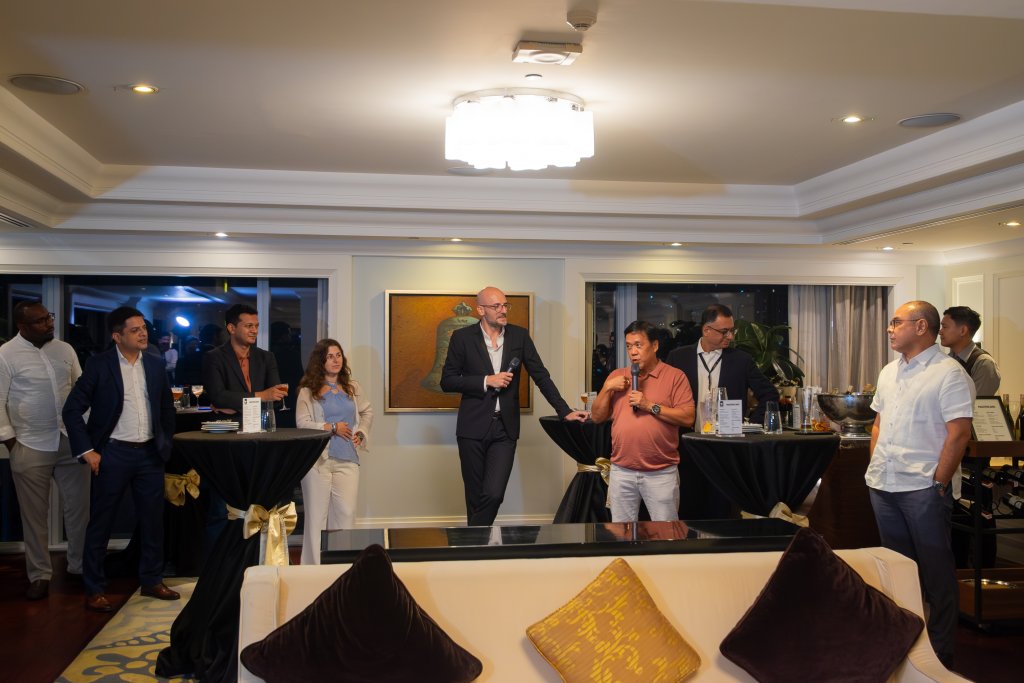 A group of eight people, both men and women, stand and socialise in a well-lit, modern living room with a chandelier. Two men at centre speak to the group. Guests stand near cocktail tables with drinks, and a gold-accented sofa is in the foreground.