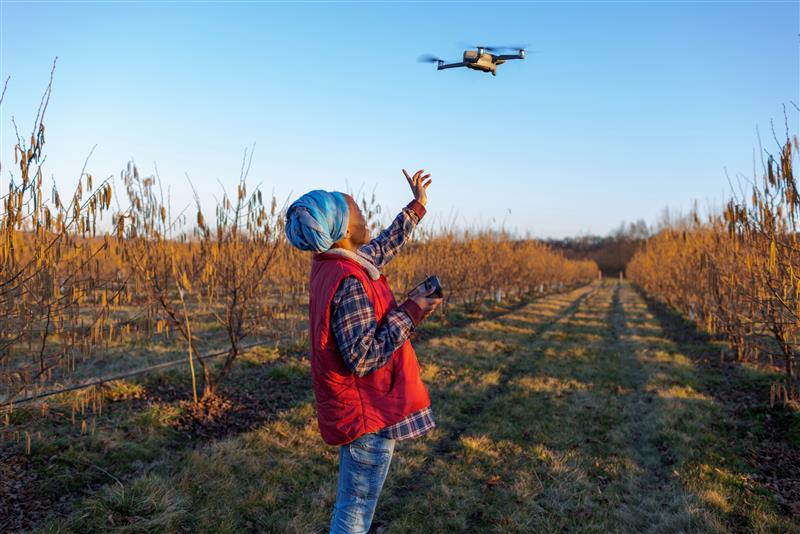 A person in a red vest and blue headscarf operates a flying drone in a field with rows of leafless trees, demonstrating AgriTech MEL for partnerships and investment under a clear blue sky. The person stands on grassy ground, reaching up toward the drone.