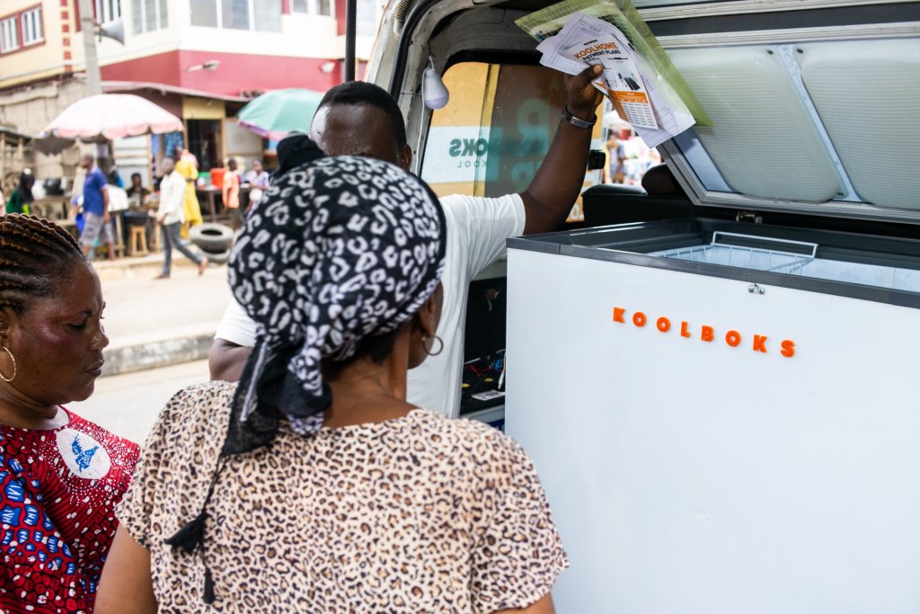Three people stand by an open white Koolboks freezer in the back of a van on a city street. One person holds papers, while two women, one in a leopard-print top and headscarf, look on. Buildings and pedestrians are visible in the background.