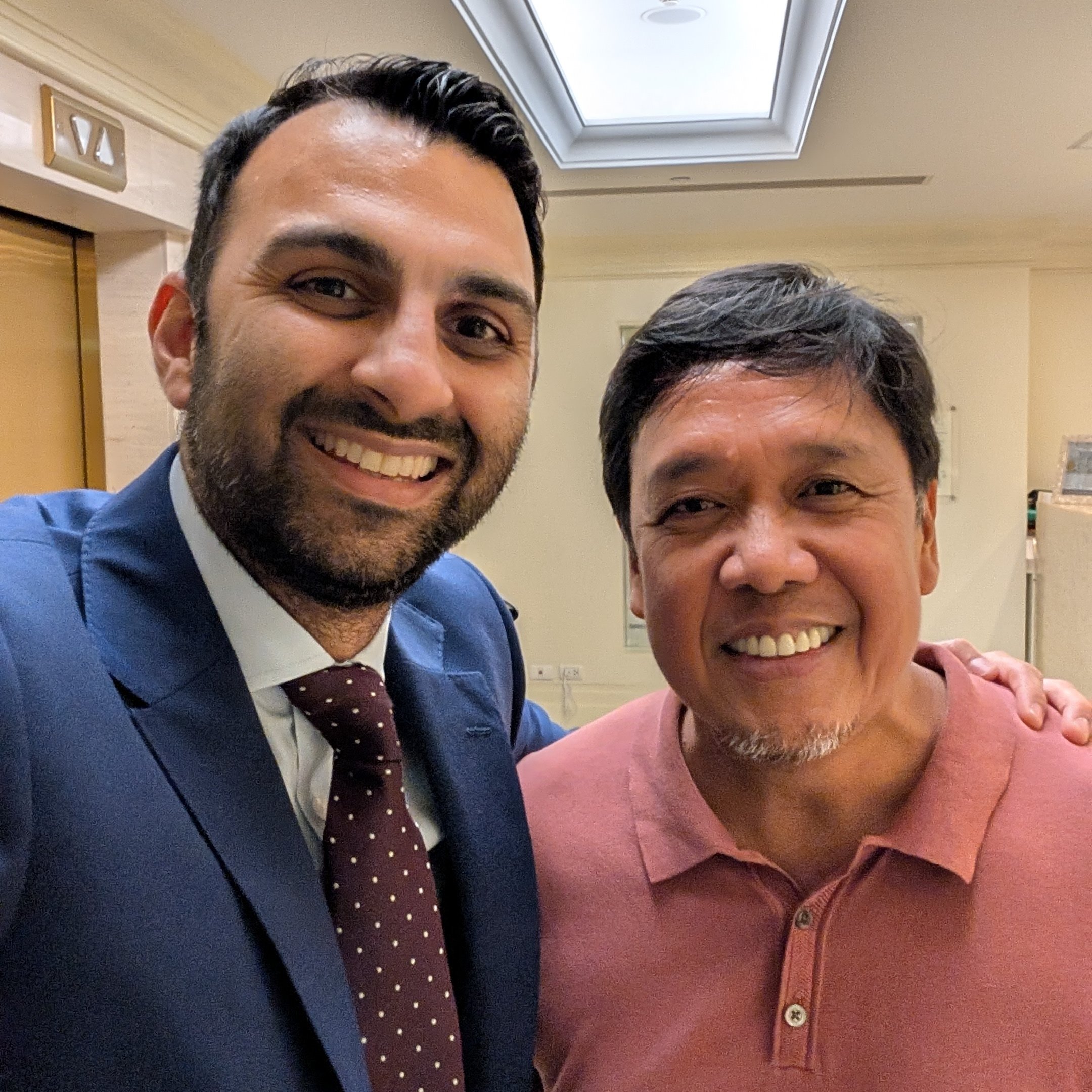 Two men smiling for a selfie indoors. The man on the left wears a navy blue suit, white shirt, and maroon polka dot tie. The man on the right wears a salmon pink polo shirt. They stand in a well-lit corridor with beige walls and a ceiling light above.