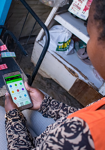A person in patterned clothing sits indoors, holding a smartphone and viewing a financial or payment app with colorful icons. Nearby are shelves holding various goods, including boxed and bagged items, in a small shop or home setting.