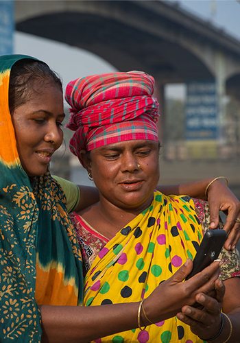 Two women in colorful traditional saris stand close together outdoors, smiling as they look at a smartphone. One has her arm around the other, who wears a red checked headscarf. A large bridge and blurry signs are visible in the background.
