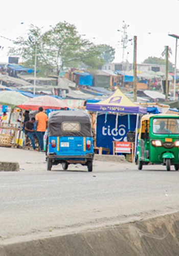 A street scene shows a blue and a green auto rickshaw driving on a paved road. In the background, people stand near colorful market stalls and umbrellas, including a prominent blue “Tigo” tent, with buildings and trees further behind.