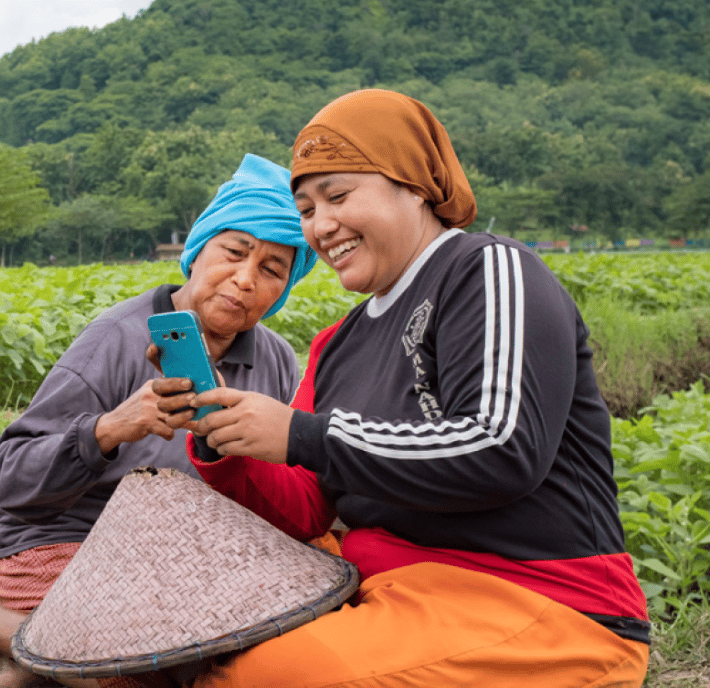 Two women in headscarves sit in a lush green field, smiling as they look at a smartphone together—a moment that reflects how technology can foster social development. A woven conical hat rests nearby, with green hills in the background.