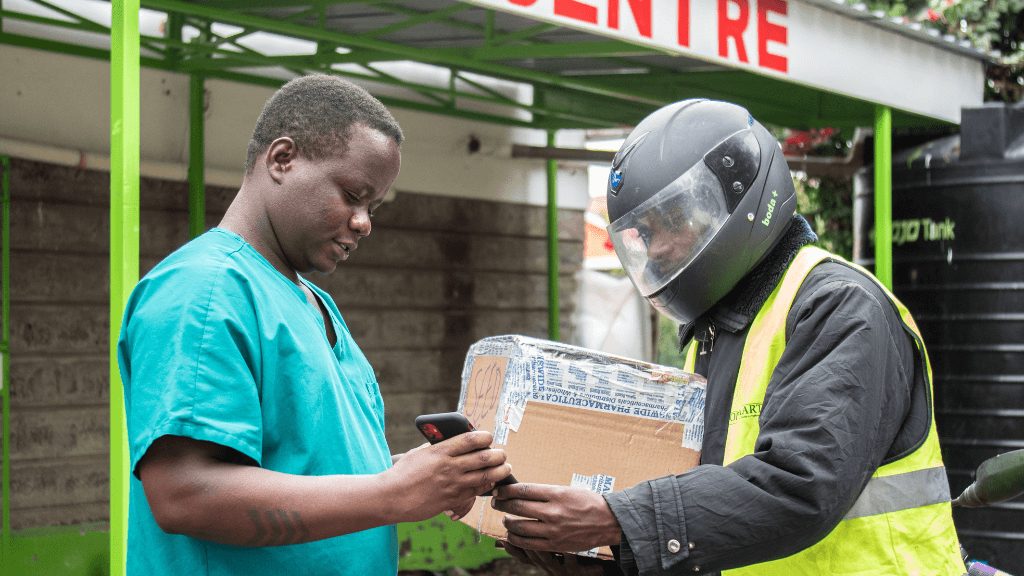 A man in teal medical scrubs demonstrates adaption as he uses a smartphone while receiving a package from a motorcycle courier in a helmet and yellow vest. They stand outside near a building with partial red text reading "CENTRE" above them.