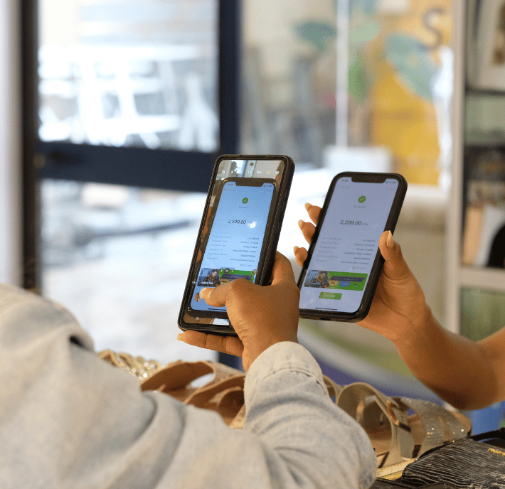 Two people hold smartphones facing each other, displaying the same mobile payment app screen. The phones show a green checkmark and payment details, enabling quick transactions as hands rest near sandals and accessories on a counter in a bright indoor setting.